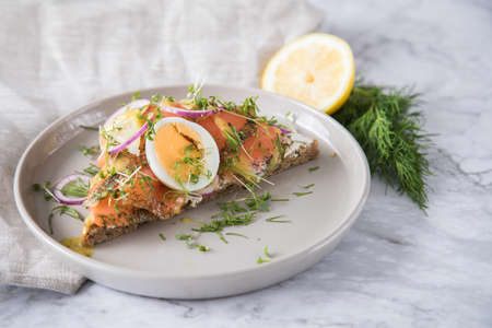Gravlax a raw, marinated graved salmon on toast bread with Scandinavian mustard sauce, dill, cress, egg, onion rings on porcelain plate linen towel and light marble backgroundの写真素材