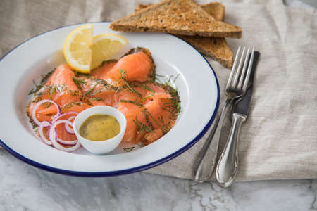Gravlax a raw, marinated graved salmon with dill on enamel plate with toast bread, Scandinavian mustard sauce, lemon, onion rings and linen towel on light marble backgroundの写真素材