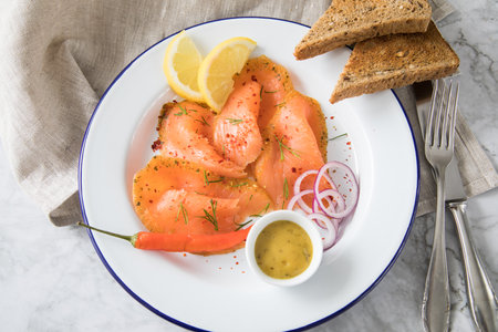 Asian style Gravlax a raw, marinated graved salmon with lemongrass and chili on enamel plate with toast bread, Scandinavian mustard sauce, dill, lemon, onion rings and linen towel on light marble backgroundの写真素材