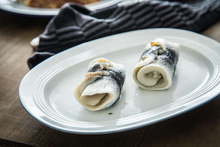 2 German Rollmops rolled pickled herring with gherkin onion filling on porcelain plate with cutlery, kitchen towel and dark wooden backgroundの写真素材