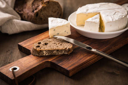 Loaf of soft blue cheese from cow milk on porcelain plate with walnut bread, knife, linen towel and dark brown wooden board as snack or dinnerの写真素材