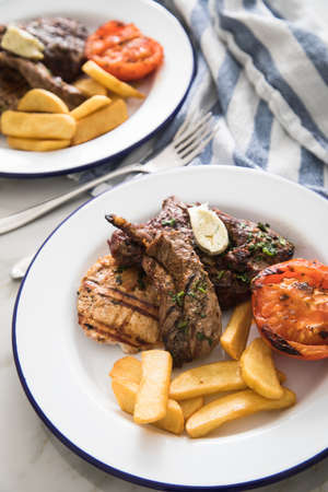 Grill platter with beef steak, chicken and lamp filet, steakhouse fries, grilled tomato, herb butter on emailed white blue plate, kitchen towel and light marble backgroundの写真素材