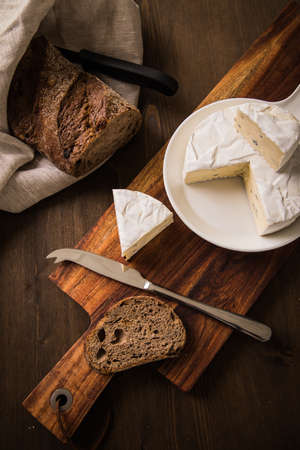 Loaf of soft blue cheese from cow milk on porcelain plate with walnut bread, knife, linen towel and dark brown wooden board as snack or dinnerの写真素材