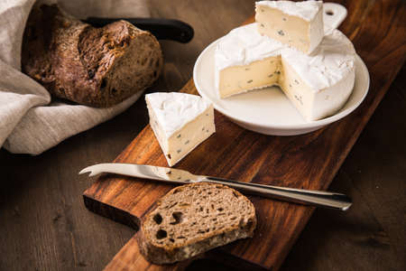 Loaf of soft blue cheese from cow milk on porcelain plate with walnut bread, knife, linen towel and dark brown wooden board as snack or dinnerの写真素材