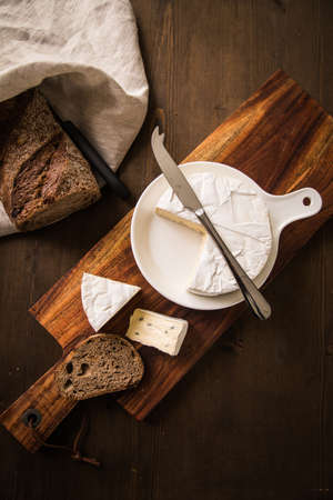 Loaf of soft blue cheese from cow milk on porcelain plate with walnut bread, knife, linen towel and dark brown wooden board as snack or dinnerの写真素材