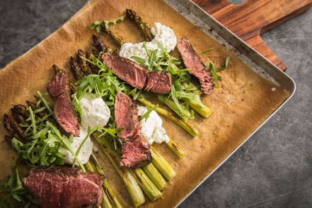 Roasted green asparagus with medium rare beef steak, rocked salad, buffalo mozzarella cheese and olive oil on baking tray, wooden board, cutlery and rustic dark stone backgroundの写真素材
