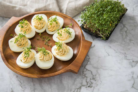 Cold deviled eggs filled with cream cheese, cress and herbs on wooden plate, linen towel and light...