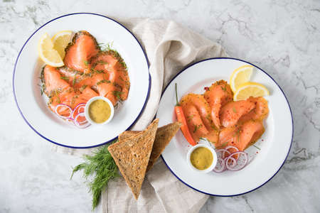 Classic and Asian style Gravlax a raw, marinated graved salmon with lemongras, chili, dill with toast bread, Scandinavian mustard sauce, dill, lemon, onion rings on enamel plate, linen towel and light marble backgroundの写真素材