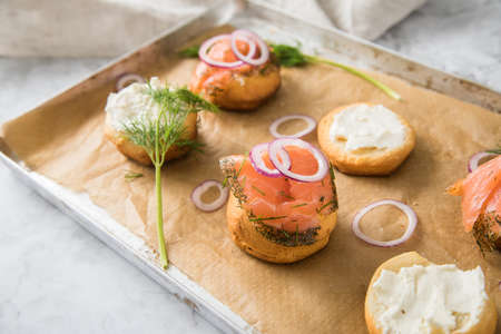 Gravlax a raw, marinated graved salmon with dill on bred roll bun with cream cheese, onion rings on tray with baking paper and light marble backgroundの写真素材