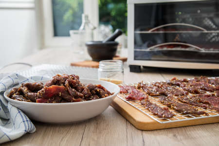 Asian style marinated raw beef strips for homemade jerky dried meat with sesame and chili on dehydrator drying tray, wooden board in light domestic kitchen for long term storageの写真素材