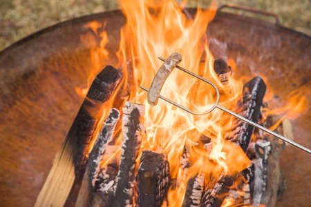 German pork Bratwurst sausage roasting on skewer over campfire of metal fire bowl in gardenの写真素材