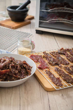 Asian style marinated raw beef strips for homemade jerky dried meat with sesame and chili on dehydrator drying tray, wooden board in light domestic kitchen for long term storageの写真素材