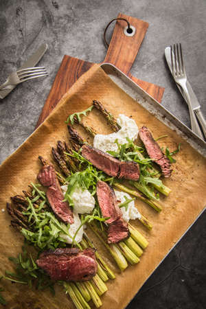 Roasted green asparagus with medium rare beef steak, rocked salad, buffalo mozzarella cheese and olive oil on baking tray, wooden board, cutlery and rustic dark stone backgroundの写真素材