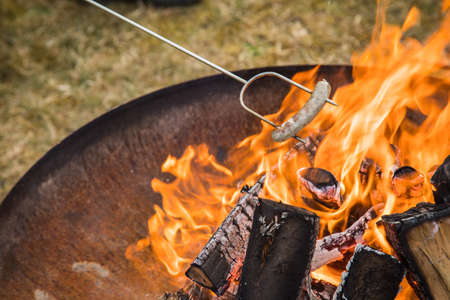 German pork Bratwurst sausage roasting on skewer over campfire of metal fire bowl in gardenの写真素材