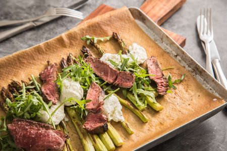 Roasted green asparagus with medium rare beef steak, rocked salad, buffalo mozzarella cheese and olive oil on baking tray, wooden board, cutlery and rustic dark stone backgroundの写真素材
