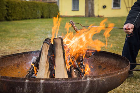 Man roasting German pork Bratwurst sausage on skewer over campfire of metal fire bowl in gardenの写真素材