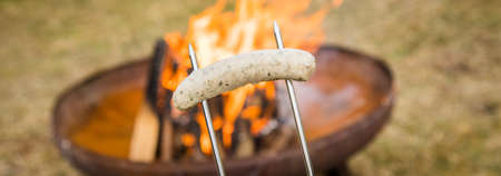 German pork Bratwurst sausage roasting on skewer over campfire of metal fire bowl in gardenの写真素材