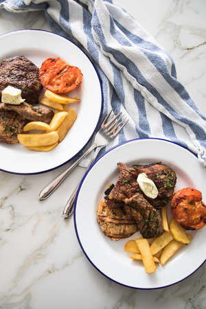 Grill platter with beef steak, chicken and lamp filet, steakhouse fries, grilled tomato, herb butter on emailed white blue plate, kitchen towel and light marble backgroundの写真素材
