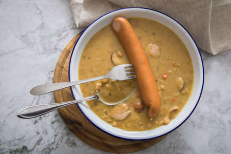 Hearty German yellow split pea soup stew with Bockwurst sausages in enamel dish on wooden board and light marble backgroundの写真素材