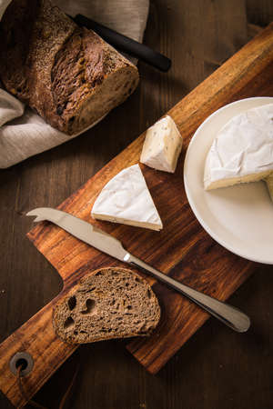 Loaf of soft blue cheese from cow milk on porcelain plate with walnut bread, knife, linen towel and dark brown wooden board as snack or dinnerの写真素材