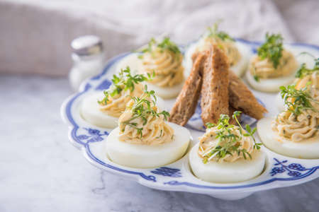 Cold deviled eggs filled with cream cheese, cress and herbs on blue white porcelain platter, linen...