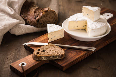 Loaf of soft blue cheese from cow milk on porcelain plate with walnut bread, knife, linen towel and dark brown wooden board as snack or dinnerの写真素材
