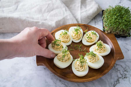 Woman with cold deviled eggs filled with cream cheese, cress and herbs on wooden plate, linen towel and light marble background for eastern, buffet, appetizer or snackの写真素材