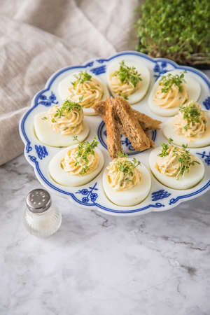 Cold deviled eggs filled with cream cheese, cress and herbs on blue white porcelain platter, linen towel and light marble background with toast bread and salt shakerの写真素材