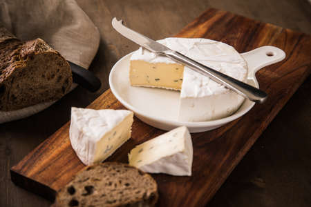 Loaf of soft blue cheese from cow milk on porcelain plate with walnut bread, knife, linen towel and dark brown wooden board as snack or dinnerの写真素材