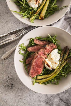Roasted green asparagus with medium rare beef steak, rocked salad, buffalo mozzarella cheese and balsamic on baking tray, wooden board, cutlery and rustic dark stone backgroundの写真素材