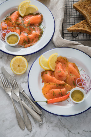 Classic and Asian style Gravlax a raw, marinated graved salmon with lemongras, chili, dill with toast bread, Scandinavian mustard sauce, dill, lemon, onion rings on enamel plate, linen towel and light marble backgroundの写真素材