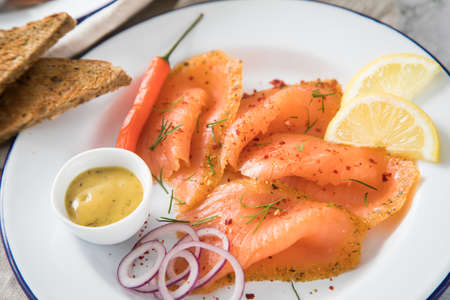Asian style Gravlax a raw, marinated graved salmon with lemongrass and chili on enamel plate with toast bread, Scandinavian mustard sauce, dill, lemon, onion rings and linen towel on light marble backgroundの写真素材