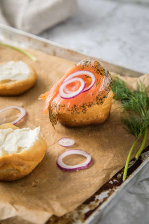Gravlax a raw, marinated graved salmon with dill on bred roll bun with cream cheese, onion rings on tray with baking paper and light marble backgroundの写真素材