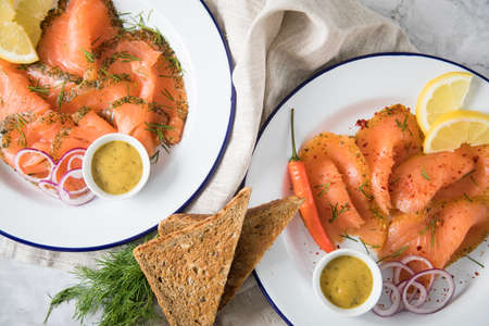 Classic and Asian style Gravlax a raw, marinated graved salmon with lemongras, chili, dill with toast bread, Scandinavian mustard sauce, dill, lemon, onion rings on enamel plate, linen towel and light marble backgroundの写真素材