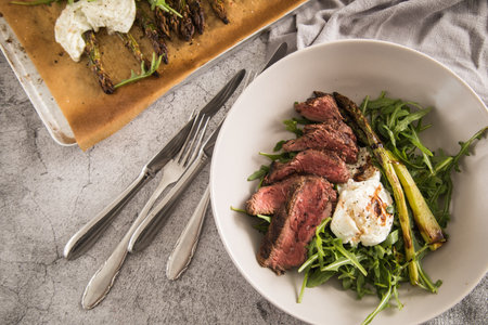 Roasted green asparagus with medium rare beef steak, rocked salad, buffalo mozzarella cheese and balsamic in bowl and baking tray, cutlery and rustic stone backgroundの写真素材