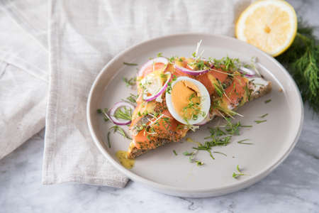 Gravlax a raw, marinated graved salmon on toast bread with Scandinavian mustard sauce, dill, cress, egg, onion rings on porcelain plate linen towel and light marble backgroundの写真素材