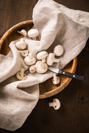 Fresh raw white Champignon mushrooms on linen towel with kitchen knife, wooden bowl and dark brown moody backgroundの写真素材