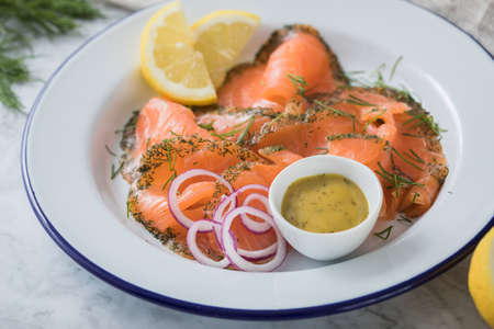 Gravlax a raw, marinated graved salmon with dill on enamel plate with Scandinavian mustard sauce, lemon, onion rings and linen towel on light marble backgroundの写真素材