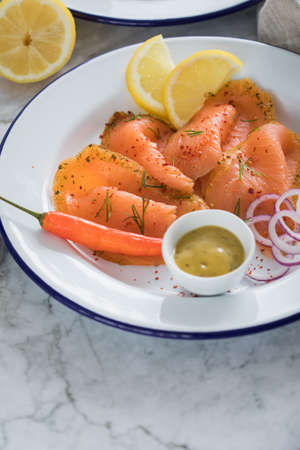 Asian style Gravlax a raw, marinated graved salmon with lemongrass and chili on enamel plate with toast bread, Scandinavian mustard sauce, dill, lemon, onion rings and linen towel on light marble backgroundの写真素材