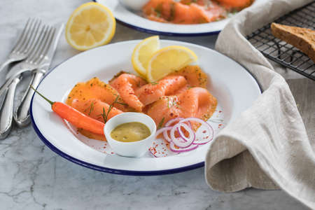 Asian style Gravlax a raw, marinated graved salmon with lemongrass and chili on enamel plate with toast bread, Scandinavian mustard sauce, dill, lemon, onion rings and linen towel on light marble backgroundの写真素材