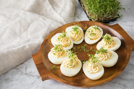 Cold deviled eggs filled with cream cheese, cress and herbs on wooden plate, linen towel and light marble background for eastern, buffet, appetizer or snackの写真素材
