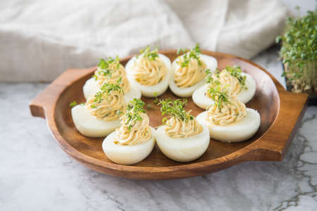 Cold deviled eggs filled with cream cheese, cress and herbs on wooden plate, linen towel and light marble background for eastern, buffet, appetizer or snackの写真素材