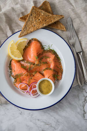 Gravlax a raw, marinated graved salmon with dill on enamel plate with toast bread, Scandinavian mustard sauce, lemon, onion rings and linen towel on light marble backgroundの写真素材