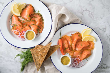 Classic and Asian style Gravlax a raw, marinated graved salmon with lemongras, chili, dill with toast bread, Scandinavian mustard sauce, dill, lemon, onion rings on enamel plate, linen towel and light marble backgroundの写真素材