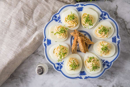 Cold deviled eggs filled with cream cheese, cress and herbs on blue white porcelain platter, linen towel and light marble background with toast bread and salt shakerの写真素材