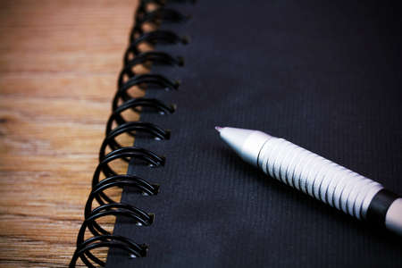 Black spiral bound notebook with pen on wooden background. Retro filter applied.