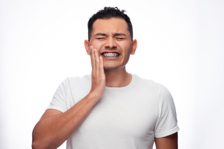 Hispanic man in white shirt and braces grimaces in pain on his face and holds his face by his orthodontic treatment brackets, one personの写真素材
