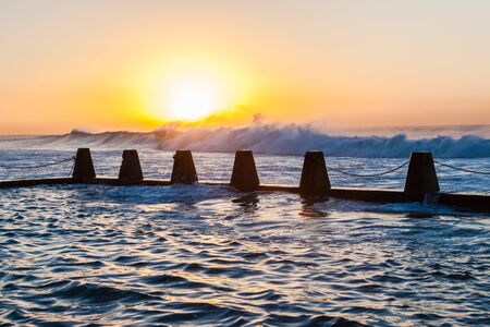 Morning sunrise with large ocean wave swells pounding crashing sea water into reef shallows and concrete beach tidal poolの写真素材