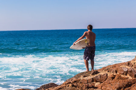 Surfer jumping off rocks to enter blue ocean to surf wavesのeditorial素材