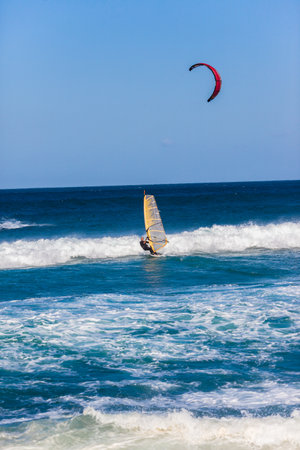 Windsurfer rides ocean waves towards the beach with kite surfers nearby 